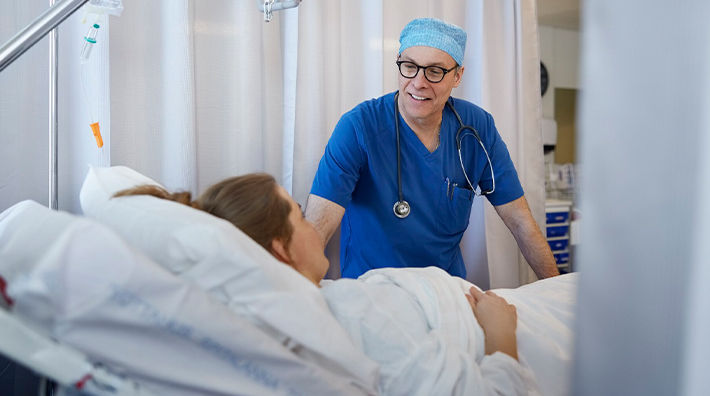A doctor wearing blue scrubs and glasses talks to a woman in a hospital bed.